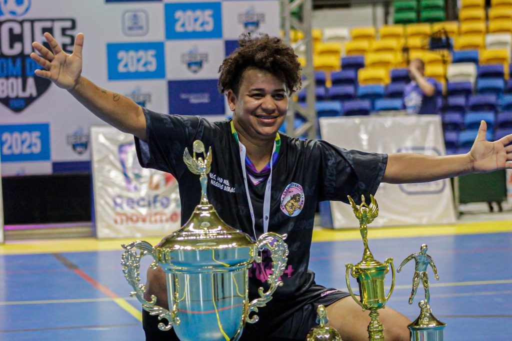 A imagem mostra Jack Mowatt, uma pessoa jovem ajoelhada em uma quadra esportiva coberta, sorrindo amplamente e abrindo os braços em sinal de comemoração. À sua frente, há três troféus metálicos de tamanhos diferentes, todos brilhantes e ornamentados — o maior tem alças curvas e uma tampa com uma pequena figura alada no topo. A pessoa veste um uniforme esportivo preto com detalhes lilás e rosa, e um crachá pendurado no pescoço. No uniforme, há um logotipo circular com o desenho de um felino e algumas palavras parcialmente legíveis. Ao fundo, vê-se arquibancadas com assentos coloridos em amarelo, azul e verde, além de banners com inscrições como “Recife”, “2025” e “Movimento”.