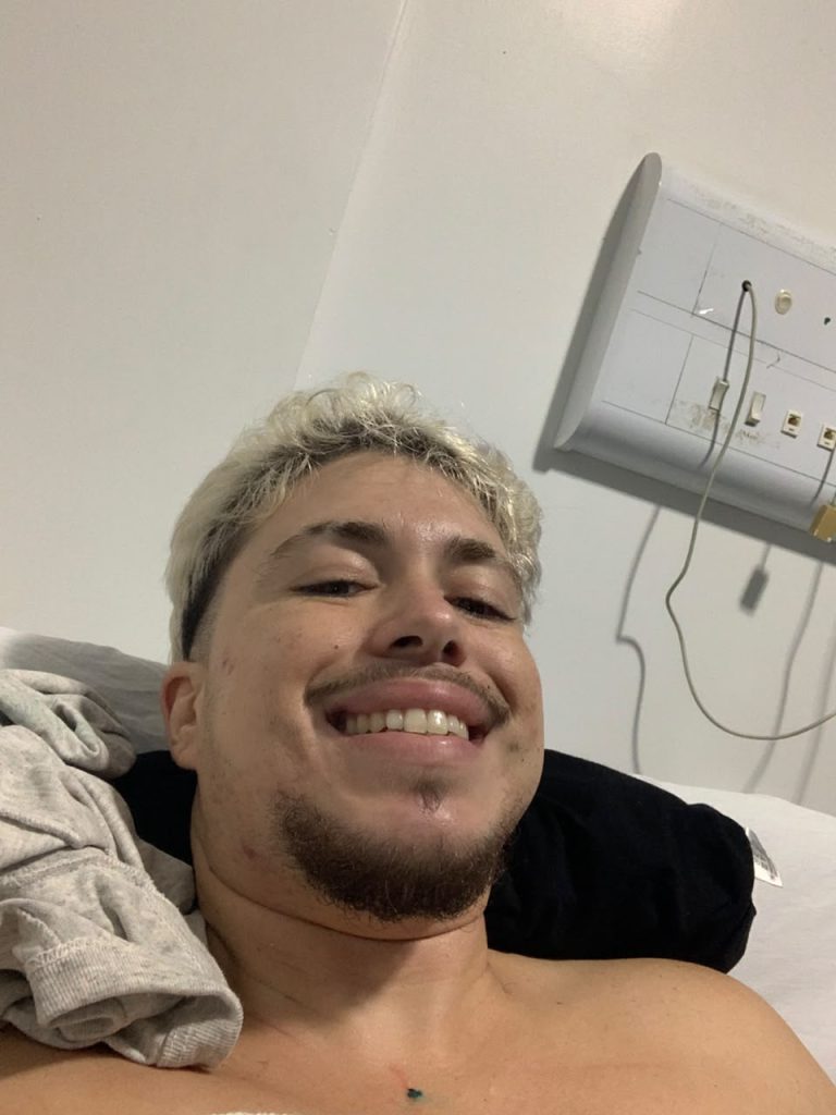A imagem mostra uma pessoa jovem deitada em uma cama, sorrindo para a câmera em um ambiente de aparência simples e clara. Essa pessoa tem pele clara, cabelo curto e loiro, barba rala e bigode. Está sem camisa e parece relaxada, com expressão tranquila e alegre. Ao lado da cabeça há uma camiseta cinza dobrada e um travesseiro preto. No fundo, vê-se uma parede branca com um painel elétrico fixado, contendo tomadas e cabos conectados — o que dá a impressão de um quarto ou ambiente hospitalar.