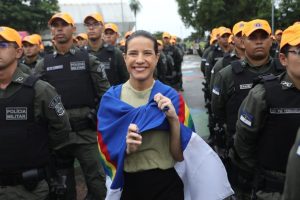 Na foto, vemos uma cena ao ar livre, em um espaço pavimentado cercado por árvores. Um grupo de policiais militares está alinhado em formação. Eles vestem uniformes verde-escuros, coletes pretos com a inscrição “POLÍCIA MILITAR” e bonés laranja bem chamativos. No centro da imagem, à frente dos policiais, está a governadora de Pernambuco Raquel Lyra envolta em uma bandeira colorida, com faixas em azul, branco, vermelho, amarelo e verde. Essa figura se destaca porque contrasta com a uniformidade dos policiais. A cena transmite a ideia de um encontro ou evento público, possivelmente uma cerimônia ou manifestação, em que há interação entre autoridades e civis.