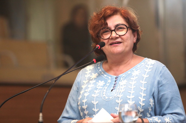 A imagem mostra Cida Pedrosa, mulher de meia-idade com cabelos curtos castanhos e óculos escuros, falando ao microfone em um ambiente de conferência. Ela veste uma blusa azul-clara bordada com desenhos brancos e segura um papel enquanto sorri de forma tranquila e confiante. À sua frente há um copo de água, e o fundo está desfocado, sugerindo um espaço formal com outras pessoas presentes.