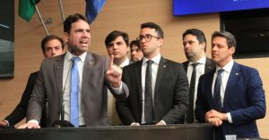 A foto mostra um grupo de homens de terno numa tribuna. Um deles está falando ao microfone e gesticulando com o dedo levantado, enquanto os demais observam com expressões sérias. O cenário sugere uma coletiva de imprensa, discurso ou pronunciamento político.