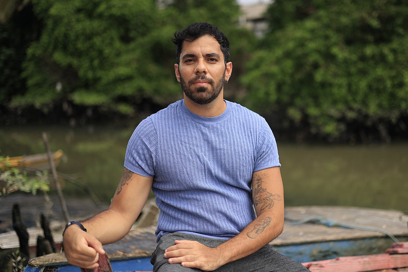 A imagem mostra Célio Rocha, um homem de pele morena e barba curta, sentado ao ar livre sobre uma superfície de madeira, possivelmente um barco ou píer. Ele veste uma camisa azul-clara de mangas curtas e calça cinza, exibindo tatuagens nos dois braços. O fundo é composto por vegetação verde densa e um rio ou canal de água barrenta, sugerindo um ambiente natural e tranquilo. Célio olha diretamente para a câmera com expressão serena e postura relaxada.