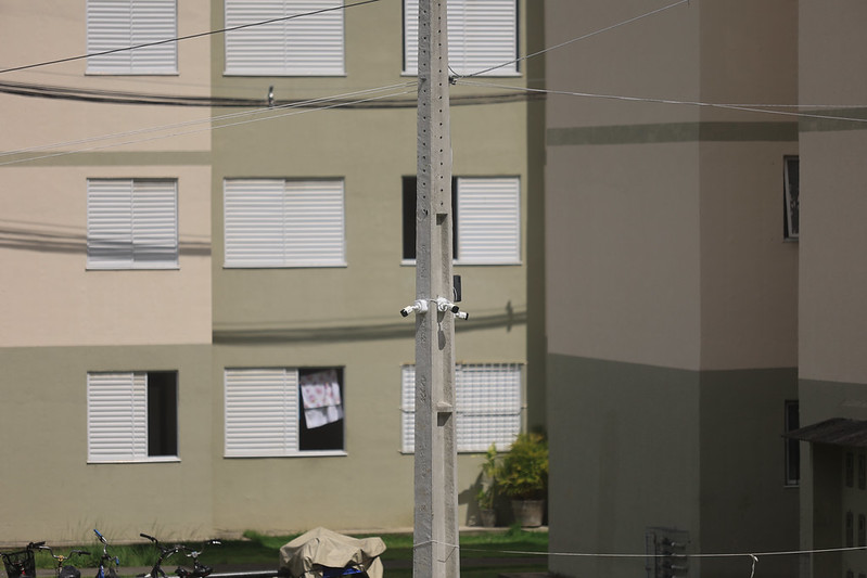 A imagem mostra um poste de concreto no centro, com câmeras de vigilância instaladas em volta dele, apontando para diferentes direções. Ao fundo, há um prédio residencial de vários andares, pintado em tons de bege e verde-oliva, com janelas de venezianas brancas, algumas abertas. Em uma das janelas, há um pano estendido ao sol. Na parte inferior, vê-se um gramado com bicicletas e um objeto coberto, possivelmente uma moto.