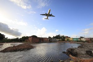 Na fotografia, um avião comercial aparece em voo baixo, ocupando o centro superior da cena contra um céu azul com nuvens brancas iluminadas pela luz suave do sol. Logo abaixo, vê-se um terreno irregular e parcialmente alagado, onde a água reflete o céu e está delimitada por uma tela de segurança laranja. Ao fundo, surgem casas simples de tijolo e concreto, algumas em construção ou sem acabamento, compondo um cenário urbano periférico. A vegetação é esparsa e se mistura com estruturas modestas, reforçando a sensação de contraste. O avião, imponente e próximo, domina a paisagem e transmite a ideia de aproximação para pouso. Já o ambiente terrestre revela o cotidiano de uma comunidade que convive diretamente com o tráfego aéreo. Essa justaposição entre a imponência tecnológica e a simplicidade residencial cria uma atmosfera de tensão e convivência singular.