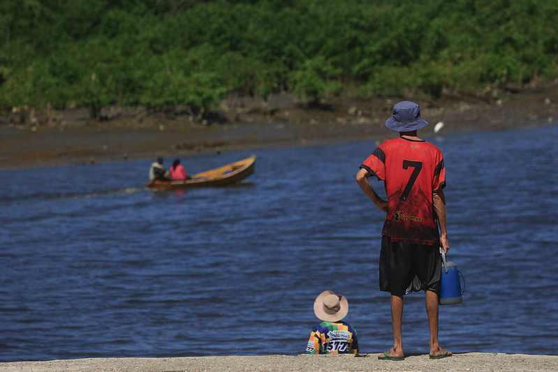 A imagem mostra uma cena tranquila à beira de um rio, onde um homem e uma criança observam um pequeno barco que navega lentamente pela água. O homem está de pé, descalço, usando uma camiseta vermelha e preta com o número 7 nas costas, um chapéu azul e segurando um galão azul na mão direita. Ao seu lado, a criança está sentada, vestindo uma camiseta colorida com números e letras e um chapéu de aba larga. No fundo, o barco de madeira leva duas pessoas — uma de camisa cinza e outra de camisa rosa — enquanto a margem oposta é coberta por vegetação verde e densa, provavelmente um manguezal. A cena transmite calma e simplicidade, como um momento cotidiano de contemplação entre o adulto e a criança diante do movimento sereno do rio.