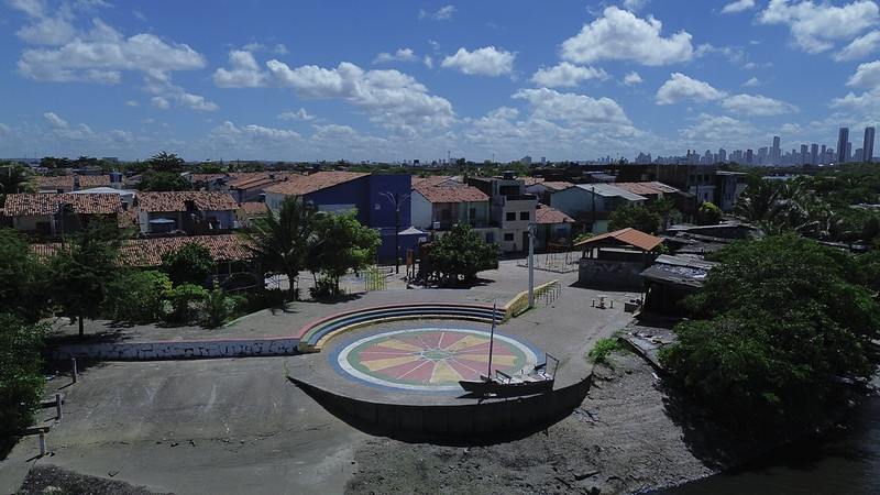 A imagem mostra uma vista aérea da Ilha de Deus, uma comunidade ribeirinha localizada no Recife. Em primeiro plano, há uma praça circular com um mosaico colorido no chão, formado por triângulos em tons de vermelho, amarelo, verde e azul, lembrando uma flor ou um sol. No centro da praça, ergue-se um mastro de bandeira, e ao redor há degraus concêntricos que formam uma espécie de anfiteatro. À esquerda e ao fundo, vê-se um conjunto de casas simples com telhados de barro e paredes pintadas em cores variadas — azul, branco, bege — cercadas por árvores e vegetação. O céu está azul e repleto de nuvens brancas, e ao longe, na linha do horizonte, aparece o perfil moderno da cidade do Recife, com seus prédios altos contrastando com o ambiente mais tradicional e tranquilo da comunidade.