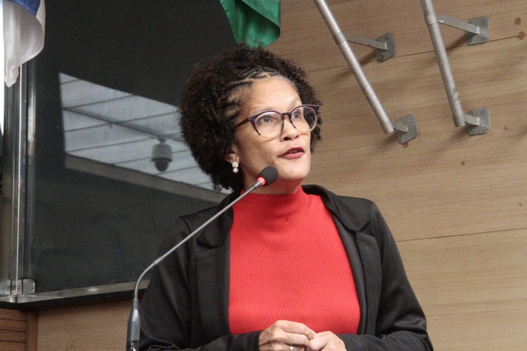 A imagem mostra Jô Cavalcanti, mulher negra de meia-idade, com cabelo crespo curto e óculos de armação escura, falando ao microfone em um ambiente formal, como uma sala de reuniões ou plenário. Ela veste uma blusa vermelha de gola alta e um blazer preto, transmitindo uma aparência elegante e profissional. O fundo tem paredes de madeira clara, partes de bandeiras e estruturas metálicas, sugerindo um espaço institucional. Sua expressão é atenta e firme, indicando que está discursando ou apresentando algo com convicção.