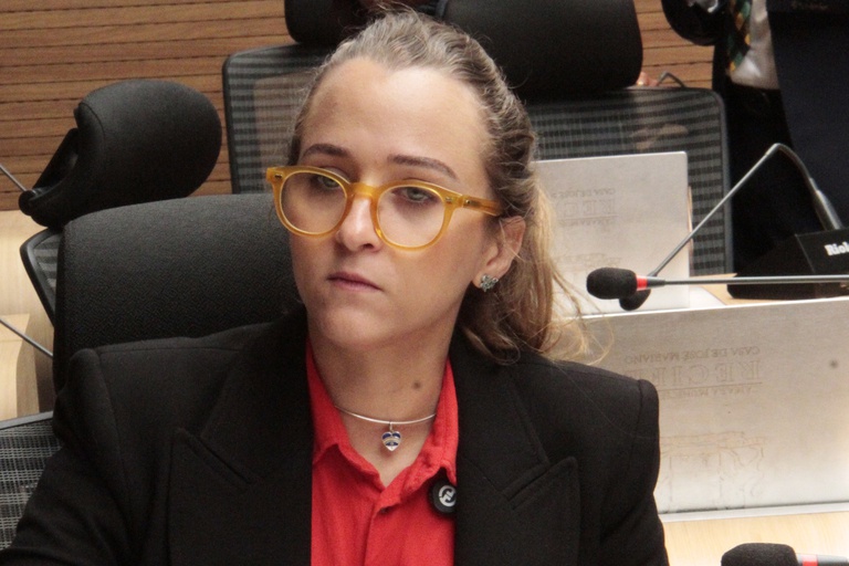 A imagem mostra Kari Santos, mulher jovem de pele clara e cabelos loiros presos, usando óculos grandes de armação amarela. Ela veste uma camisa vermelha e um blazer preto, com um colar prateado e um pingente pequeno em formato de coração. Kari está sentada em um ambiente formal, possivelmente uma sala de reuniões ou plenário, com microfones e cadeiras de escritório ao redor. Sua expressão é séria e concentrada, sugerindo atenção ao que está sendo discutido.
