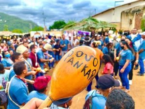A foto mostra uma grande reunião de pessoas ao ar livre, aparentemente em um evento comunitário ou cultural. Em primeiro plano, uma pessoa segura um instrumento de percussão com a inscrição “Povo Pankará”, enquanto várias pessoas participam da celebração ao fundo.