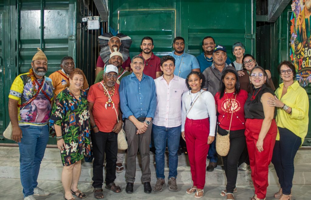 A foto mostra um grupo de cerca de 15 pessoas posando juntas para uma foto em um ambiente interno. Algumas pessoas usam roupas e acessórios tradicionais e indígenas, enquanto outras estão vestidas de forma casual. O grupo está sorrindo, transmitindo um clima de confraternização, diversidade cultural e celebração coletiva.