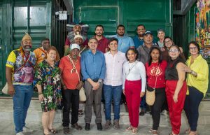 A foto mostra um grupo de cerca de 15 pessoas posando juntas para uma foto em um ambiente interno. Algumas pessoas usam roupas e acessórios tradicionais e indígenas, enquanto outras estão vestidas de forma casual. O grupo está sorrindo, transmitindo um clima de confraternização, diversidade cultural e celebração coletiva.