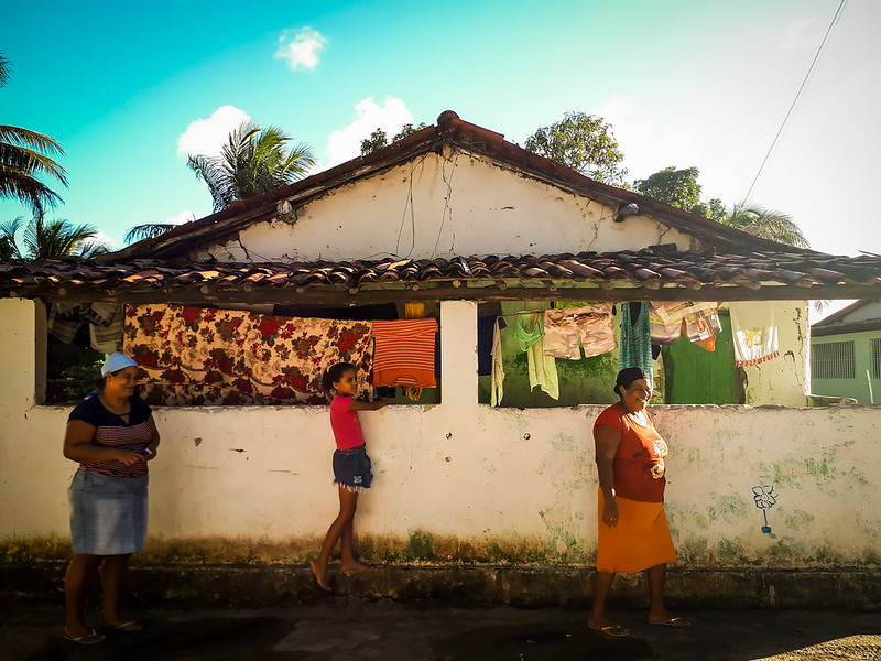 A imagem mostra uma casa simples, de um andar, com telhado de barro e paredes brancas um pouco desgastadas. Na frente, há roupas coloridas penduradas para secar — vermelhas, verdes, listradas — e um grande tecido floral cobrindo parte do muro. Três pessoas estão na calçada: uma mulher à esquerda com saia jeans e camiseta listrada, uma menina no centro encostada no muro, e outra mulher à direita vestindo blusa vermelha e saia laranja. O céu está azul e claro, com algumas nuvens e palmeiras ao fundo, sugerindo um clima quente e tranquilo de região tropical.