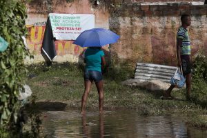 A imagem mostra duas pessoas em uma área alagada e cheia de lama, próximas a um muro desgastado com cartazes antigos. Uma delas, uma mulher de blusa e guarda-chuva azul que caminha no alagamento, enquanto a outra, um homem de blusa listrada e bermuda carrega uma bolsa infantil.