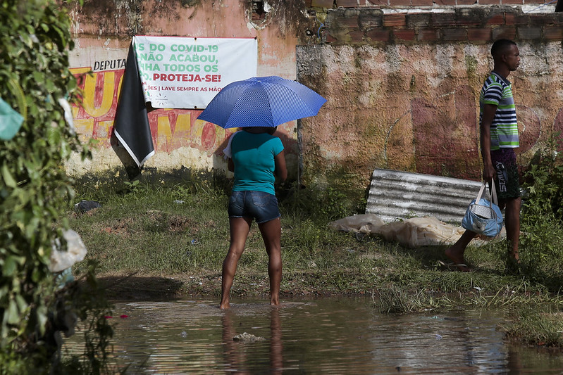 A imagem mostra duas pessoas em uma área alagada e cheia de lama, próximas a um muro desgastado com cartazes antigos. Uma delas, uma mulher de blusa e guarda-chuva azul que caminha no alagamento, enquanto a outra, um homem de blusa listrada e bermuda carrega uma bolsa infantil.