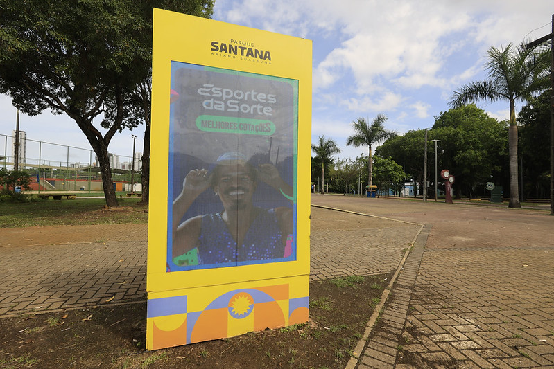 A imagem mostra um totem digital amarelo instalado em uma área aberta do Parque Santana – Ariano Suassuna, em Recife. No visor do totem há um anúncio da marca “Esportes da Sorte”, com o texto “Melhores cotações” e a foto de uma pessoa sorridente. O entorno apresenta árvores, palmeiras e uma quadra esportiva cercada, com piso de tijolos e céu parcialmente nublado — um ambiente urbano e bem cuidado, típico de um parque público.