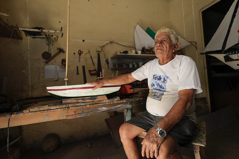 A foto mostra o pescador conhecido como Carpinteiro, um senhor de idade sentado em seu ateliê de trabalho. Ele veste uma camiseta branca com o texto “Festa de São Pedro” e uma imagem de uma jangada, além de shorts escuros e um relógio prateado no pulso. Carpinteiro está sentado em um banco de madeira, com uma expressão serena e concentrada, enquanto apoia a mão sobre um modelo de barco em construção sobre a bancada. O ambiente ao redor é simples e cheio de ferramentas, tintas e materiais de artesanato, revelando o espaço onde ele cria suas miniaturas de embarcações.