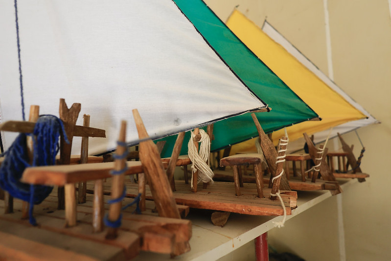 A foto mostra pequenos modelos de jangadas artesanais dispostos sobre uma prateleira. Cada jangada é feita de madeira rústica, com velas triangulares coloridas — branca, verde e amarela — presas por cordas e fios. Os detalhes incluem bancos, mastros e amarras feitos à mão, com fios azuis e brancos enrolados nas estruturas. O fundo é simples, com uma parede bege, destacando o trabalho manual e o cuidado nos detalhes dessas miniaturas.