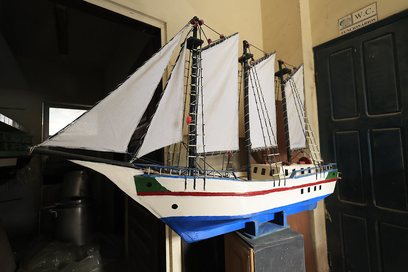 A imagem mostra um modelo de navio à vela exposto em um ambiente interno. O navio tem três mastros com velas brancas e cordas pretas que formam a armação. O casco é pintado de branco, com uma faixa vermelha e detalhes verdes e azuis na parte inferior. Ele está apoiado sobre um suporte azul. Ao fundo, há uma porta escura com uma placa indicando “W.C. Funcionários” e uma abertura que dá para outro cômodo. A foto destaca o trabalho artesanal e o cuidado nos detalhes do modelo.