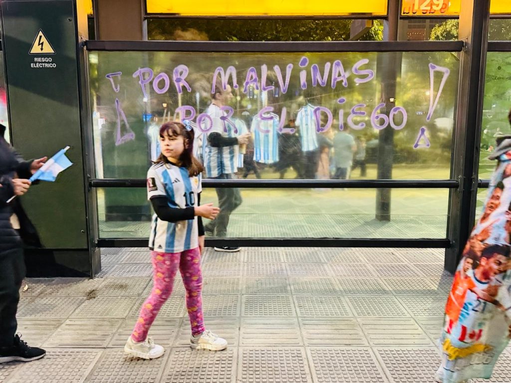 Esta fotografia de Sebás Miquel captura um momento de forte identidade argentina. No centro, uma menina veste a camisa número 10 da seleção — símbolo eterno de Diego Maradona — e caminha diante de um abrigo de ônibus onde se lê, em letras roxas: “Por Malvinas, por el Diego!”. À esquerda, alguém segura uma pequena bandeira da Argentina; à direita, parte de uma manta exibe rostos de jogadores. O reflexo no vidro mostra outras pessoas com o mesmo uniforme, sugerindo uma celebração coletiva, talvez após um jogo.