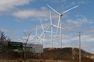 A foto mostra um parque eólico instalado em uma região de clima seco e terreno acidentado. No centro e ao fundo da imagem, há vários aerogeradores brancos — torres altas com três pás cada — alinhados sobre as colinas, girando lentamente com o vento. O céu está parcialmente nublado, com manchas de azul entre as nuvens. No primeiro plano, o solo é coberto por vegetação rasteira e arbustos secos, típicos de áreas semiáridas. Há também placas de sinalização e postes com fios elétricos cruzando a cena. As placas trazem nomes como “Fundo de Desenvolvimento do Nordeste (FDNE)” e “Banco do Nordeste”, indicando que o projeto tem apoio de instituições regionais.