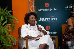 Rosane Borges, uma mulher preta, cabelos crespos acima do ombro, com um vestido branco com detalhes em renda, fala ao microfone sentada em cadeira de madeira. Ao fundo, parede laranja e painel verde com o nome “Livraria do Jardim Espaço Plural” e plantas decorativas.