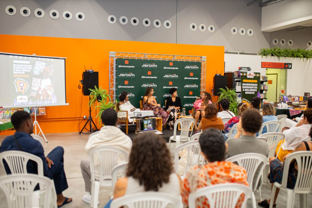 A imagem mostra um debate em uma livraria com quatro pessoas sentadas em um palco diante de um público em cadeiras brancas. O espaço tem paredes laranjas, plantas decorativas e um painel verde com o nome “Jardim”. À esquerda, um telão exibe o cartaz “Imaginários Emergentes e Mulheres Negras” com ilustrações de uma mulher, um tigre e o símbolo feminista. O mesmo cartaz aparece à direita em um suporte próximo a livros, reforçando o tema literário e cultural do evento.