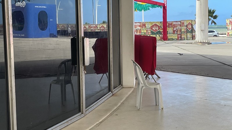 A foto mostra uma área externa com piso de concreto liso e uma parede de vidro refletindo o ambiente. Em frente ao vidro há uma cadeira plástica branca, posicionada próxima ao canto do prédio. Dentro, o reflexo revela outra cadeira escura. Ao fundo, aparecem duas estruturas vermelhas cobertas por tecido, provavelmente parte de um evento ou montagem. Mais distante, há decorações coloridas penduradas e, além delas, o mar e palmeiras, indicando um local praiano.