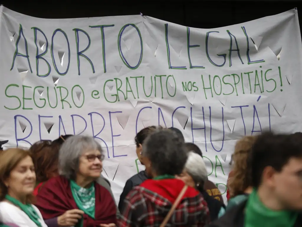 A foto mostra um grupo de pessoas reunidas diante de um grande cartaz branco com letras escritas à mão em azul e verde. O texto do cartaz diz: “Aborto legal, seguro e gratuito nos hospitais! Reabre Cachutá!”. As pessoas aparecem em primeiro plano, conversando e caminhando, muitas delas usando lenços verdes no pescoço — símbolo amplamente associado à luta feminista pela legalização do aborto na América Latina.