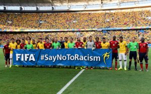 A imagem mostra jogadores de duas seleções de futebol lado a lado em campo, segurando uma grande faixa azul com os dizeres “FIFA #SayNoToRacism”. Eles estão uniformizados — uma equipe veste amarelo e azul, e a outra, vermelho e branco — e atrás há um estádio lotado de torcedores, majoritariamente vestidos de amarelo. É uma cena de união e apoio à campanha contra o racismo, promovendo respeito e igualdade no esporte.
