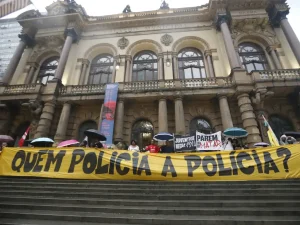 A manifestação retratada ocorre diante do Teatro Municipal de São Paulo, onde pessoas sob guarda-chuvas exibem faixas com mensagens contra a violência policial, como “Quem policia a polícia?”, “Juventude negra viva” e “Parem de nos matar”. A cena, marcada pela arquitetura imponente e pelo clima chuvoso, expressa um ato público de denúncia e resistência, destacando o apelo por justiça e controle das ações policiais no Brasil.