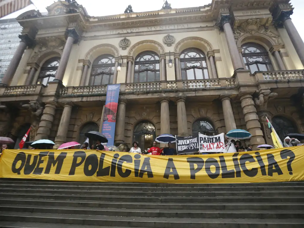 A manifestação retratada ocorre diante do Teatro Municipal de São Paulo, onde pessoas sob guarda-chuvas exibem faixas com mensagens contra a violência policial, como “Quem policia a polícia?”, “Juventude negra viva” e “Parem de nos matar”. A cena, marcada pela arquitetura imponente e pelo clima chuvoso, expressa um ato público de denúncia e resistência, destacando o apelo por justiça e controle das ações policiais no Brasil.