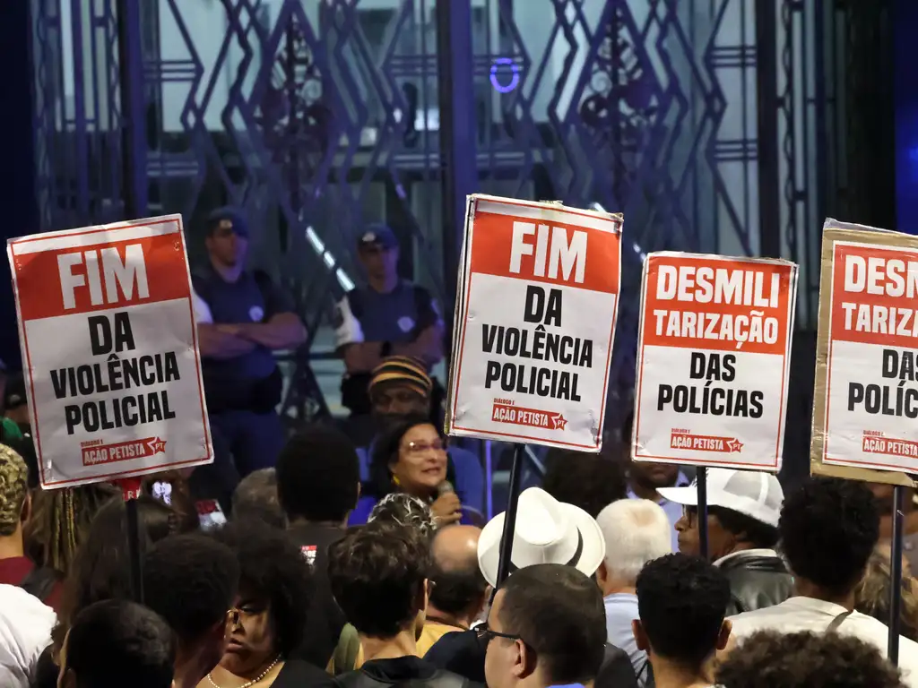 A foto mostra uma manifestação com grande número de pessoas reunidas, segurando cartazes que pedem o fim da violência policial e a desmilitarização das polícias. Os cartazes, em vermelho e branco, trazem o selo “Diálogos da Ação Petista”, enquanto ao fundo alguns policiais observam a cena próximos a um portão metálico. A imagem expressa um momento de mobilização social e reivindicação por mudanças na segurança pública e no modo como as forças policiais atuam no país.