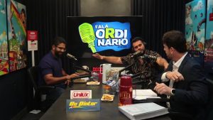 A imagem mostra três homens participando de um podcast chamado “Fala Ordinário”. Eles estão sentados em torno de uma mesa com microfones, copos, papéis e alguns petiscos. No fundo, há uma tela com o logotipo do programa — um microfone amarelo e o nome em letras grandes sobre um fundo azul. O ambiente é colorido, com um ar de aparente descontração, com quadros vibrantes nas paredes que retratam cenas urbanas e elementos artísticos. À esquerda, há uma placa divertida que diz “Perigo: área sujeita a ataque de muricoca”. Os participantes parecem estar em meio a uma conversa animada — um deles gesticula com entusiasmo enquanto os outros escutam.