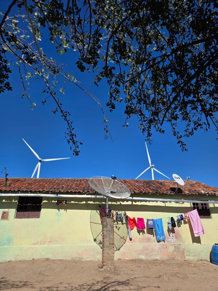 A foto mostra uma casa simples de parede amarelada e telhado de barro, típica de área rural, com roupas coloridas penduradas em um varal à frente. No quintal, há uma grande antena parabólica apoiada em um pilar de tijolos e outra menor no telhado, marcada com o logotipo “Vivo”. Ao fundo, destacam-se imensas turbinas eólicas brancas sob um céu azul intenso, sugerindo a presença de um parque de energia renovável. Galhos de uma árvore sombreiam parcialmente a cena, criando um contraste entre o cotidiano rural e os avanços tecnológicos que se integram à paisagem.