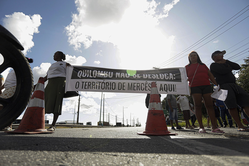 A foto mostra um grupo de pessoas em uma estrada, segurando uma faixa que diz: “Quilombo não se desloca: o território de Mercês é aqui!”. Elas estão posicionadas atrás de cones de trânsito, sob um céu claro e ensolarado. A cena transmite um ato de protesto quilombola, em defesa do direito à terra e à permanência no território tradicional da comunidade de Mercês.