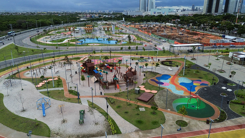 A foto aérea mostra o Parque Eduardo Campos, em Recife, com amplas áreas verdes e caminhos sinuosos que conectam diferentes espaços de lazer. No centro há um grande playground colorido com escorregadores, pontes e balanços, cercado por pisos pintados em tons vibrantes. Ao redor, vê-se árvores jovens, postes de iluminação e outras áreas recreativas, incluindo lagos artificiais e estruturas de madeira. O parque é margeado por avenidas largas e, ao fundo, surgem prédios altos e uma zona em construção sob um céu nublado, compondo um cenário urbano moderno e dinâmico.