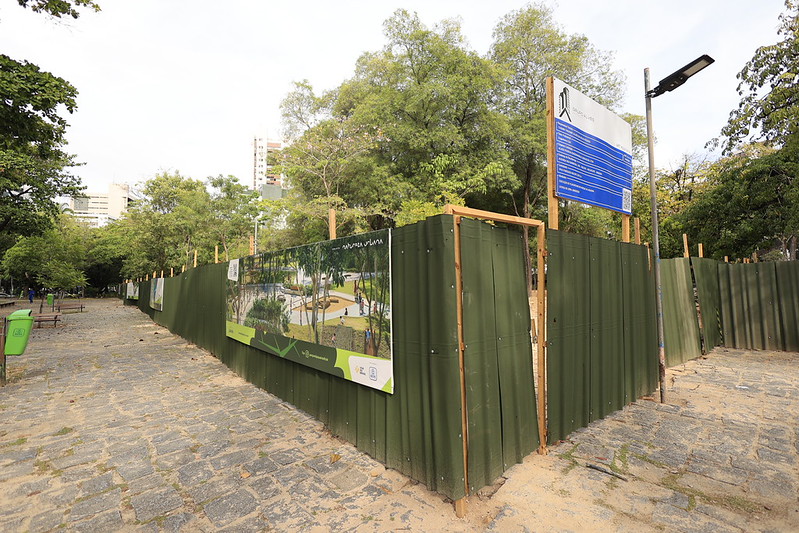 A foto mostra uma área do Parque da Jaqueira, no Recife, cercada por chapas metálicas verdes sustentadas por estruturas de madeira, indicando uma obra de revitalização em andamento. Na cerca há um painel com uma ilustração do projeto “Natureza Urbana”, mostrando caminhos, árvores e espaços de convivência, e ao lado um quadro azul com informações técnicas sobre a intervenção. O chão é de pedras irregulares, formando uma trilha entre árvores frondosas e bancos de madeira; à esquerda, uma lixeira verde reforça o cuidado ambiental do local. Ao fundo, surgem prédios altos entre as copas das árvores, compondo a paisagem urbana do Recife.