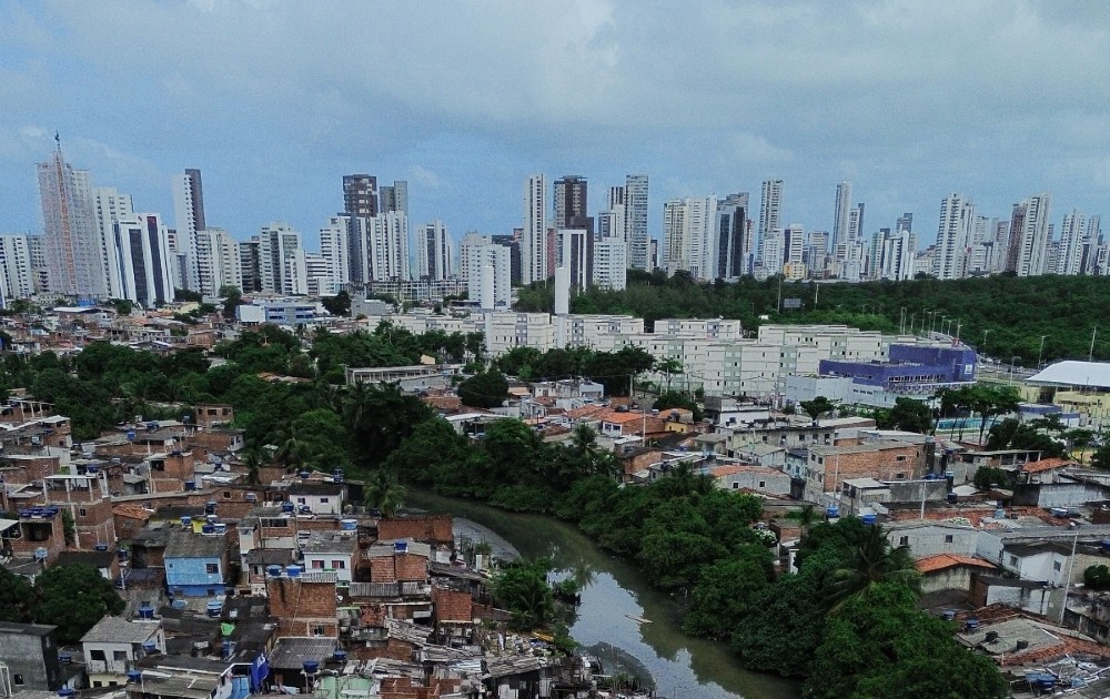 Vista aérea da Comunidade do Bode, no Recife, com casas de alvenaria às margens de um braço de rio, conjuntos habitacionais ao centro e prédios da Zona Sul ao fundo