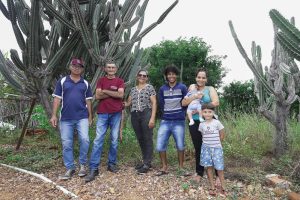 A foto mostra seis pessoas em pé sobre um terreno pedregoso, cercadas por grandes cactos e vegetação verde. Elas estão alinhadas e sorrindo para a câmera, em um ambiente que parece rural e semiárido. À esquerda, há três adultos — dois homens e uma mulher — e à direita, um homem jovem, uma mulher segurando um bebê e uma criança pequena à frente. Todos vestem roupas casuais, como camisetas, calças jeans e sandálias. O cenário transmite uma sensação de comunidade e conexão com a natureza.