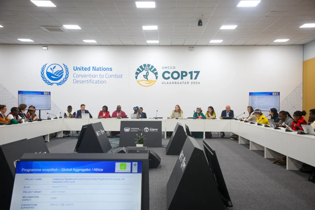 A foto mostra uma reunião oficial da Convenção das Nações Unidas de Combate à Desertificação durante a COP17, que ocorrerá em Ulaanbaatar em 2026, com várias pessoas sentadas em torno de uma mesa em formato de “U”, cada uma com microfone e computador, participando de uma discussão sobre o programa Indigenous Peoples and Local Communities Programme for Locally Led Adaptation, financiado pelo Fundo de Adaptação e implementado pelo PNUD; o ambiente é moderno, bem iluminado e decorado com os logotipos da ONU e da COP17.