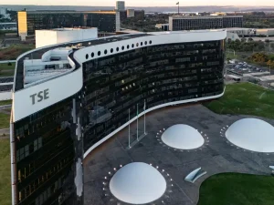 A imagem mostra o edifício moderno do Tribunal Superior Eleitoral (TSE), em Brasília. A construção tem formato curvo, fachada de vidro escuro e detalhes brancos nas extremidades superiores. Em frente ao prédio, há três estruturas brancas em forma de cúpula, dispostas sobre uma ampla área pavimentada, com pequenas luzes ao redor e mastros de bandeiras ao centro. O entorno é composto por gramados e outros prédios públicos, característicos da arquitetura monumental da capital brasileira.