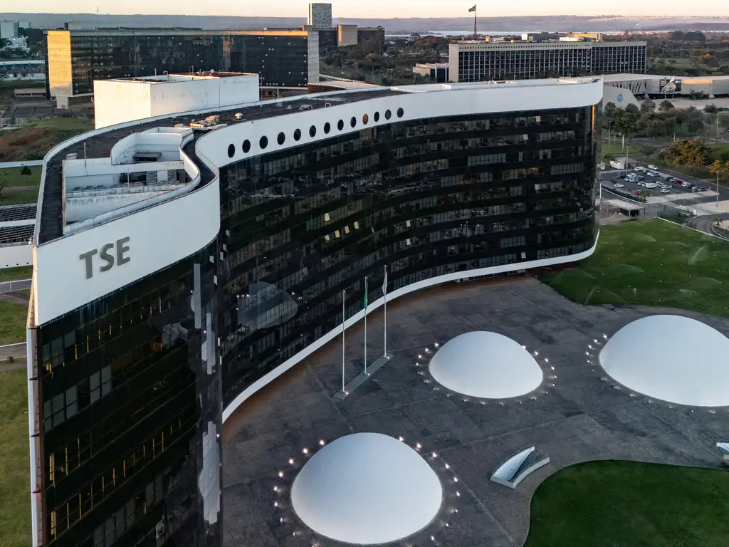 A imagem mostra o edifício moderno do Tribunal Superior Eleitoral (TSE), em Brasília. A construção tem formato curvo, fachada de vidro escuro e detalhes brancos nas extremidades superiores. Em frente ao prédio, há três estruturas brancas em forma de cúpula, dispostas sobre uma ampla área pavimentada, com pequenas luzes ao redor e mastros de bandeiras ao centro. O entorno é composto por gramados e outros prédios públicos, característicos da arquitetura monumental da capital brasileira.