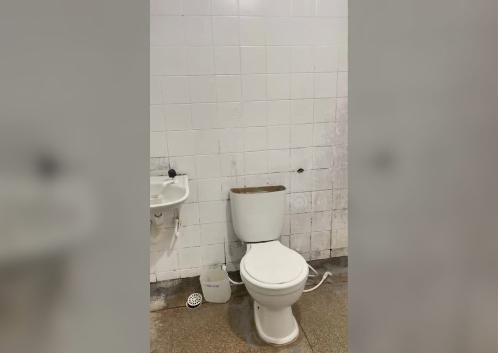 A foto mostra um pequeno banheiro simples e um pouco desgastado. No centro há um vaso sanitário branco, sem tampa no reservatório de água, deixando o interior visível. À esquerda do vaso, há uma pia branca pequena fixada na parede, com torneira e canos aparentes. O fundo é formado por uma parede de azulejos brancos, alguns escurecidos ou manchados, especialmente próximos ao chão e à pia. No piso, que parece ser de cimento ou cerâmica marrom, há um balde plástico branco com inscrições parcialmente legíveis e uma tampa de ralo circular branca caída ao lado. O ambiente aparenta estar úmido e precisar de limpeza ou manutenção.