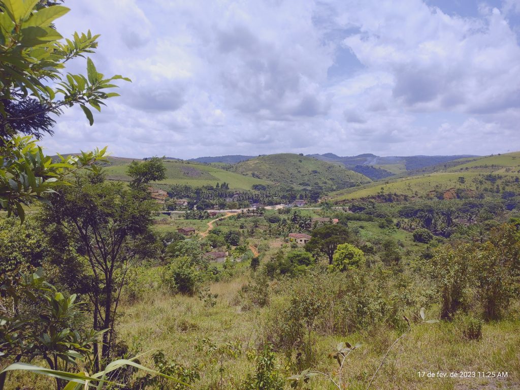 A imagem mostra uma paisagem rural vista de um ponto alto, com colinas verdes que se estendem até o horizonte sob um céu parcialmente nublado. No vale, há uma pequena comunidade com casas espalhadas e uma estrada de terra que corta o cenário, cercada por vegetação densa e árvores de diferentes tamanhos. A luz suave do fim da manhã realça os tons de verde e transmite uma sensação de tranquilidade e isolamento em meio à natureza.