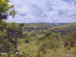 A imagem mostra uma paisagem rural vista de um ponto alto, com colinas verdes que se estendem até o horizonte sob um céu parcialmente nublado. No vale, há uma pequena comunidade com casas espalhadas e uma estrada de terra que corta o cenário, cercada por vegetação densa e árvores de diferentes tamanhos. A luz suave do fim da manhã realça os tons de verde e transmite uma sensação de tranquilidade e isolamento em meio à natureza.