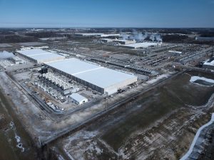 A imagem mostra um data center semelhante a grande complexo industrial visto de cima, provavelmente capturado por um drone ou avião. Há vários galpões retangulares enormes com telhados claros, cercados por instalações menores, tubulações e áreas de estacionamento. É possível ver vapor saindo de algumas partes, indicando atividade intensa — talvez processos de fabricação ou sistemas de aquecimento. O entorno é composto por terreno plano com partes cobertas de neve ou geada, estradas pavimentadas e pequenas lagoas artificiais. Ao fundo, há linhas de transmissão e outras construções industriais, sugerindo uma área dedicada à produção em larga escala.