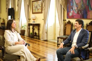 A imagem mostra Raquel Lyra, governadora de Pernambuco, e João Campos, ex-prefeito do Recife, sentados frente a frente em uma sala elegante e iluminada, conversando de forma cordial. Ela veste roupas claras e sorri, enquanto ele usa paletó azul escuro, camisa branca e jeans. O ambiente tem piso de madeira polida, móveis antigos e cortinas claras nas janelas altas; ao fundo há uma mesa com um globo decorativo e plantas, além de quadros coloridos nas paredes. A cena transmite uma atmosfera de diálogo amistoso em um espaço institucional ou histórico, com destaque para os detalhes artísticos e arquitetônicos do local.