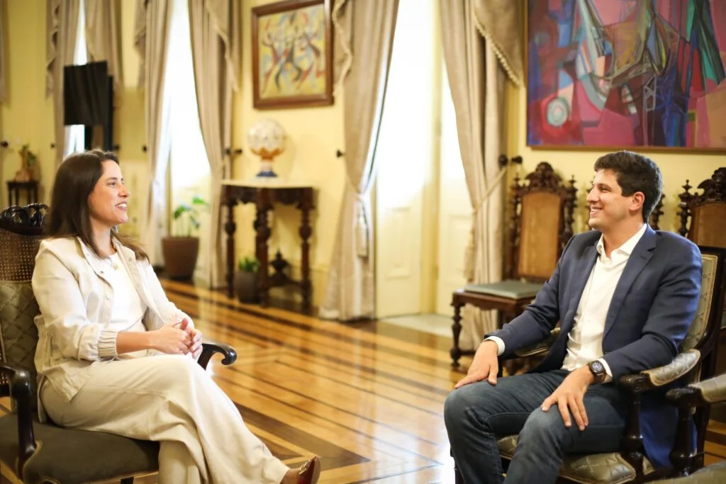 A imagem mostra Raquel Lyra, governadora de Pernambuco, e João Campos, ex-prefeito do Recife, sentados frente a frente em uma sala elegante e iluminada, conversando de forma cordial. Ela veste roupas claras e sorri, enquanto ele usa paletó azul escuro, camisa branca e jeans. O ambiente tem piso de madeira polida, móveis antigos e cortinas claras nas janelas altas; ao fundo há uma mesa com um globo decorativo e plantas, além de quadros coloridos nas paredes. A cena transmite uma atmosfera de diálogo amistoso em um espaço institucional ou histórico, com destaque para os detalhes artísticos e arquitetônicos do local.