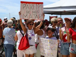 A foto mostra uma manifestação feminista em um local ensolarado, com várias pessoas reunidas segurando cartazes que denunciam a violência contra as mulheres e pedem justiça. Em destaque, uma mulher ergue um cartaz com a frase “PAREM DE NOS MATAR”, enquanto uma menina segura outro que diz “ESTAMOS AQUI PELA CRIMINALIZAÇÃO DA MISOGINIA”. O ambiente transmite união, força e resistência, com participantes de diferentes idades expressando solidariedade e exigindo igualdade de gênero.