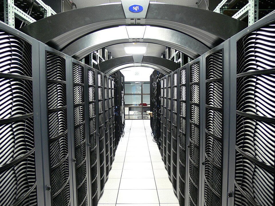 A imagem mostra o interior de um centro de dados — um ambiente técnico onde ficam fileiras de servidores organizados em racks metálicos pretos. O piso é branco e elevado, típico para permitir a passagem de cabos e ventilação. No teto, há arcos e luminárias que iluminam o corredor central. Ao fundo, vê-se uma porta de vidro que leva a outra sala. O logotipo azul “sgi” indica que o equipamento pertence à Silicon Graphics International, empresa especializada em computação de alto desempenho.